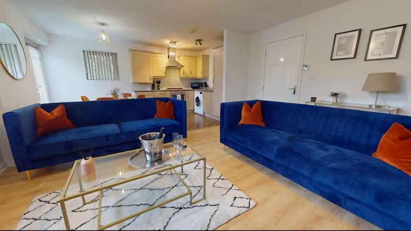 Open-plan living room with two blue sofas, orange cushions, glass coffee table and kitchen visible