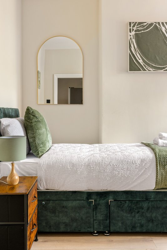 Modern bedroom with green upholstered bed, white bedding, gold-framed mirror, and wooden side table