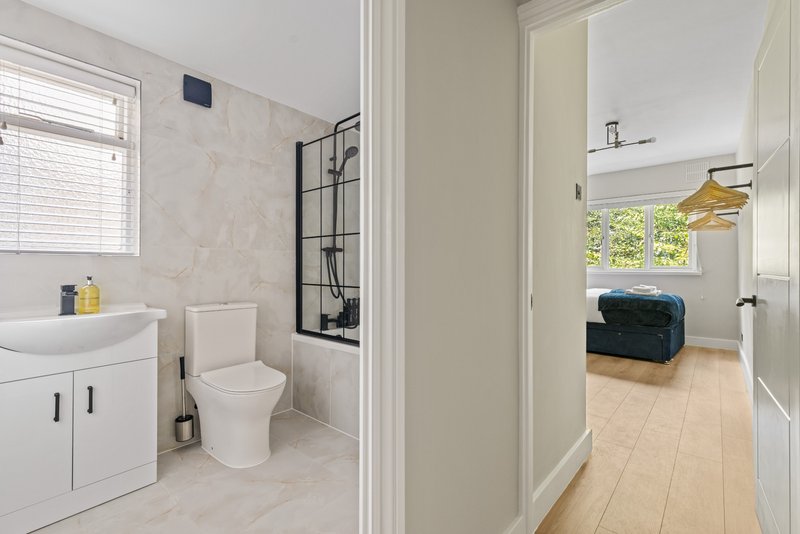 Modern bathroom with white vanity, toilet, and shower enclosure, adjacent to hallway with bedroom visible