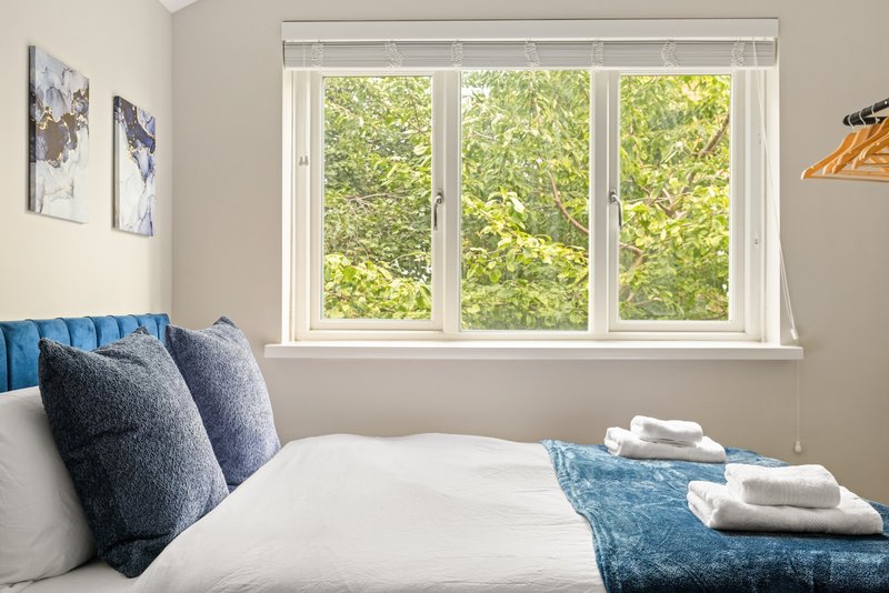 Bedroom with double bed, blue cushions, white bedding, and large window overlooking green trees