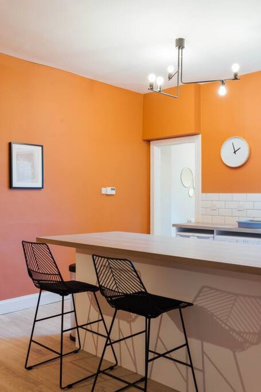 Modern kitchen with orange walls, white countertop, black bar stools and wall clock