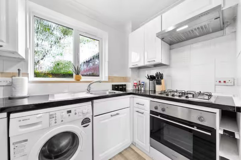 Modern white kitchen with black countertops, built-in oven, gas hob, washing machine and window overlooking garden