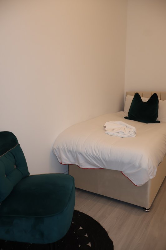 Compact bedroom with single bed, white duvet, black cushion, and teal armchair