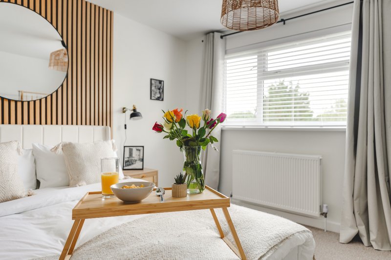 Modern bedroom with wooden bed frame, round mirror, fresh flowers on wooden table and window with grey curtains