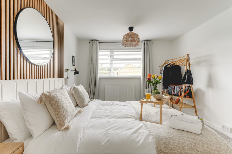 Bright modern bedroom with white bedding, round mirror, wooden accent wall and clothing rack