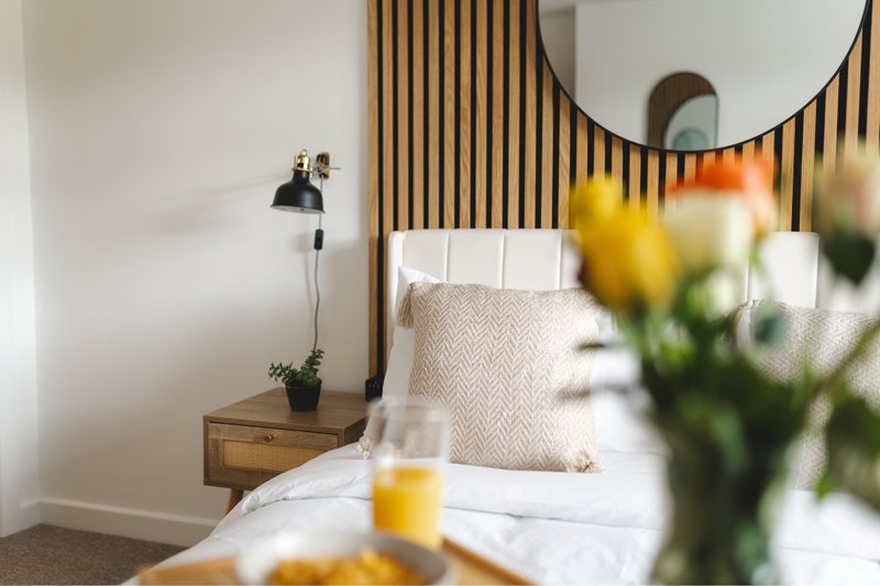Modern bedroom with double bed, wooden headboard, round mirror, bedside lamp and fresh flowers