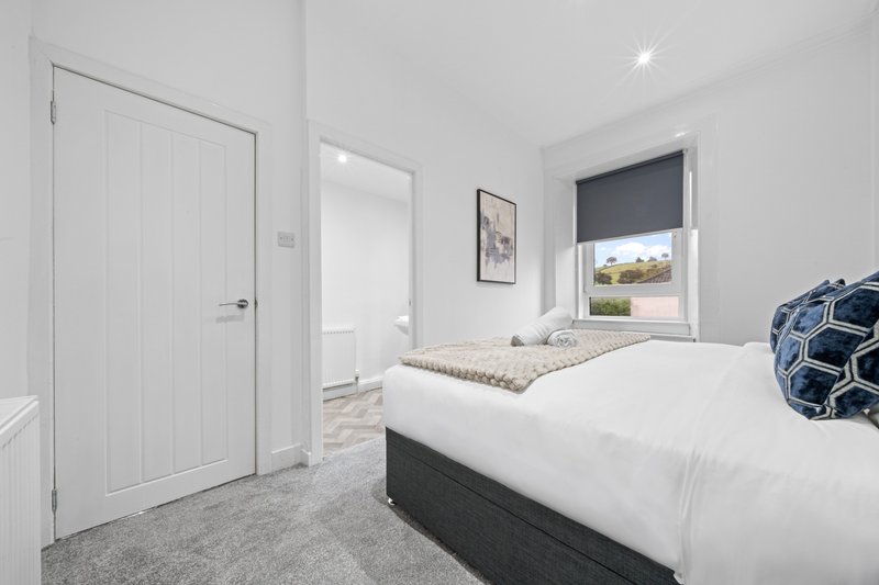 Bright modern bedroom with twin beds, white doors, window with dark blind, and grey carpet flooring.