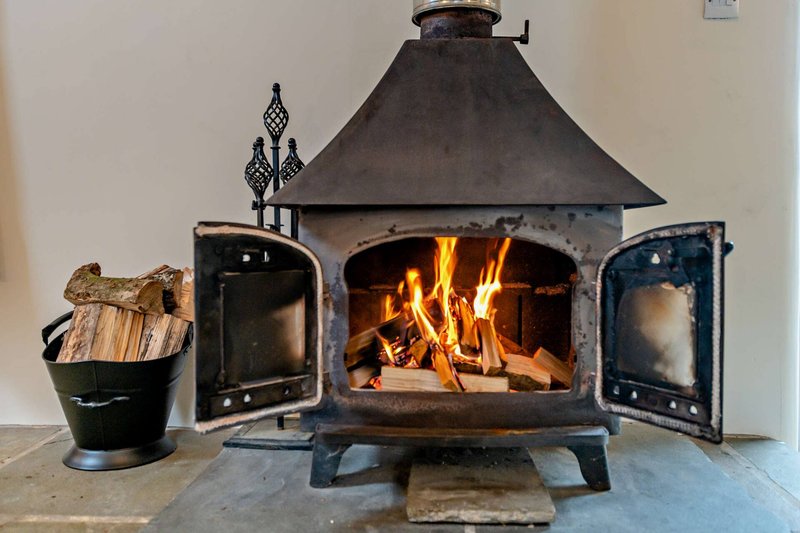Black metal wood-burning stove with lit fire, doors open, and firewood bucket beside it