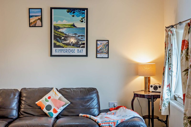 Cozy living room with dark leather sofa, colorful cushions, framed travel posters, and table lamp