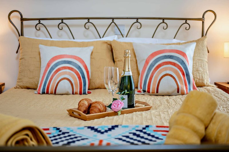 Bedroom with ornate headboard, decorative rainbow pillows, champagne bottle, glasses and fresh bread on bed