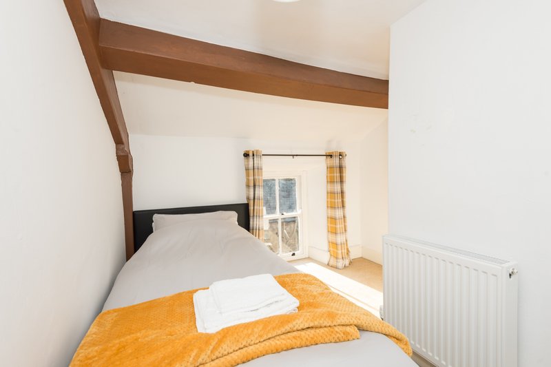 Small bedroom with single bed, orange bedding, exposed wooden beam and window with yellow curtains