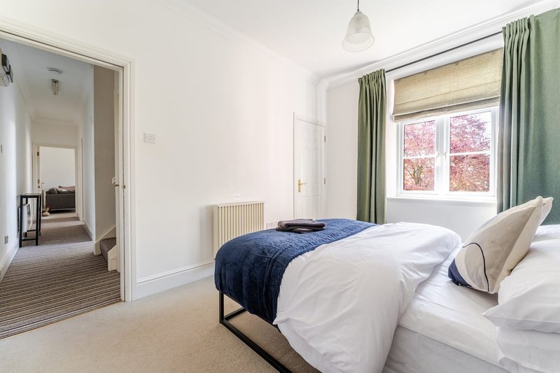 Bright bedroom with double bed, white bedding, blue throw, green curtains and window view
