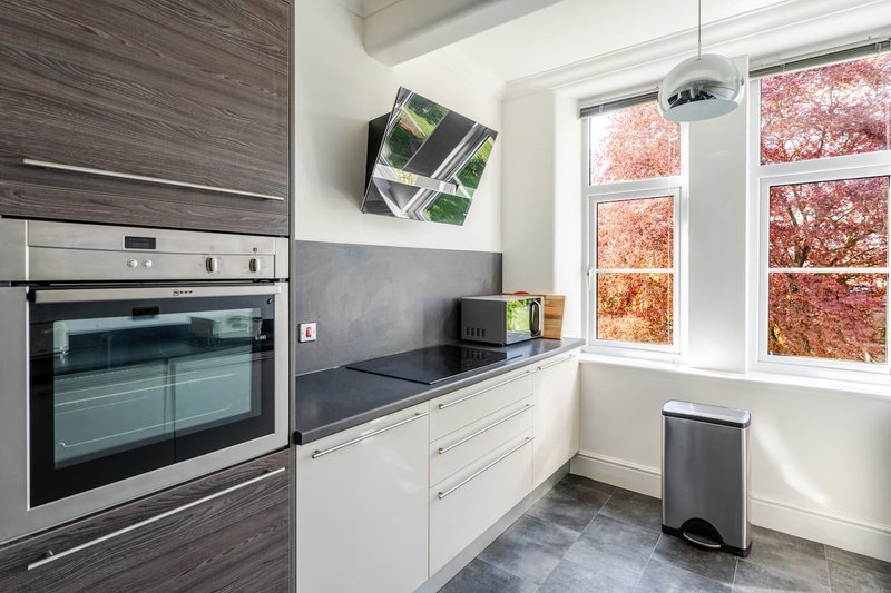 Modern kitchen with white cabinetry, black countertop, built-in oven, and window overlooking autumn trees