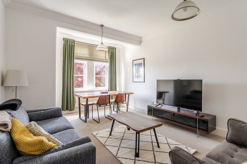 Open-plan living room with sofa, dining table, TV unit and large windows with green curtains