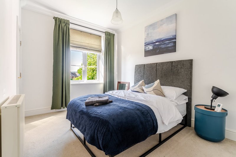 Bright bedroom with double bed, grey headboard, blue bedding, and green curtains