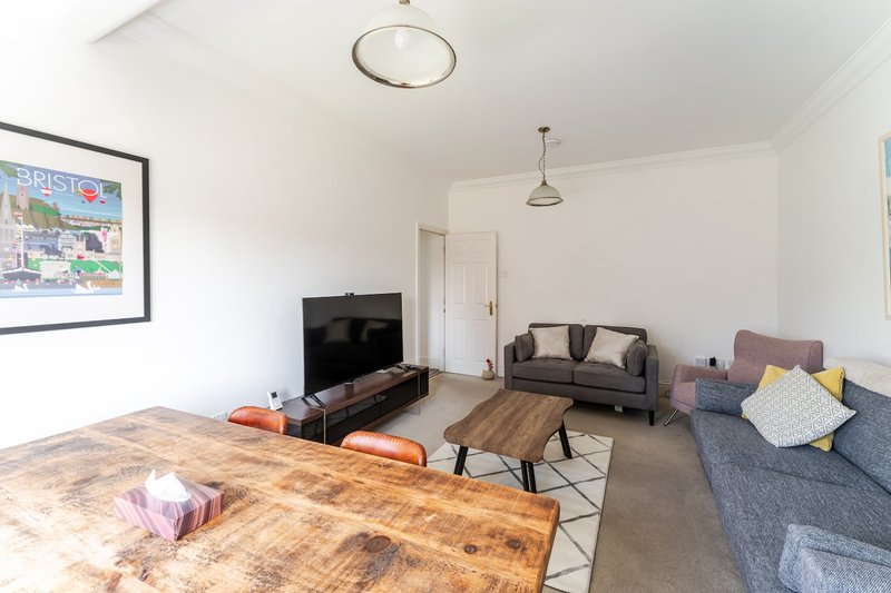 Bright open-plan living area with wooden dining table, grey sofas, TV, and framed wall art