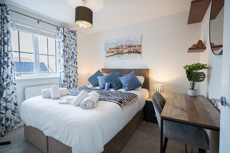 Modern bedroom with double bed, blue cushions, desk, window with patterned curtains and coastal artwork