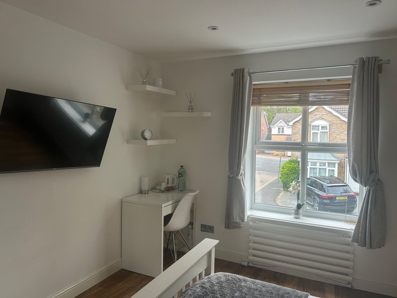 Modern bedroom with wall-mounted TV, white desk, window overlooking street, and wooden flooring