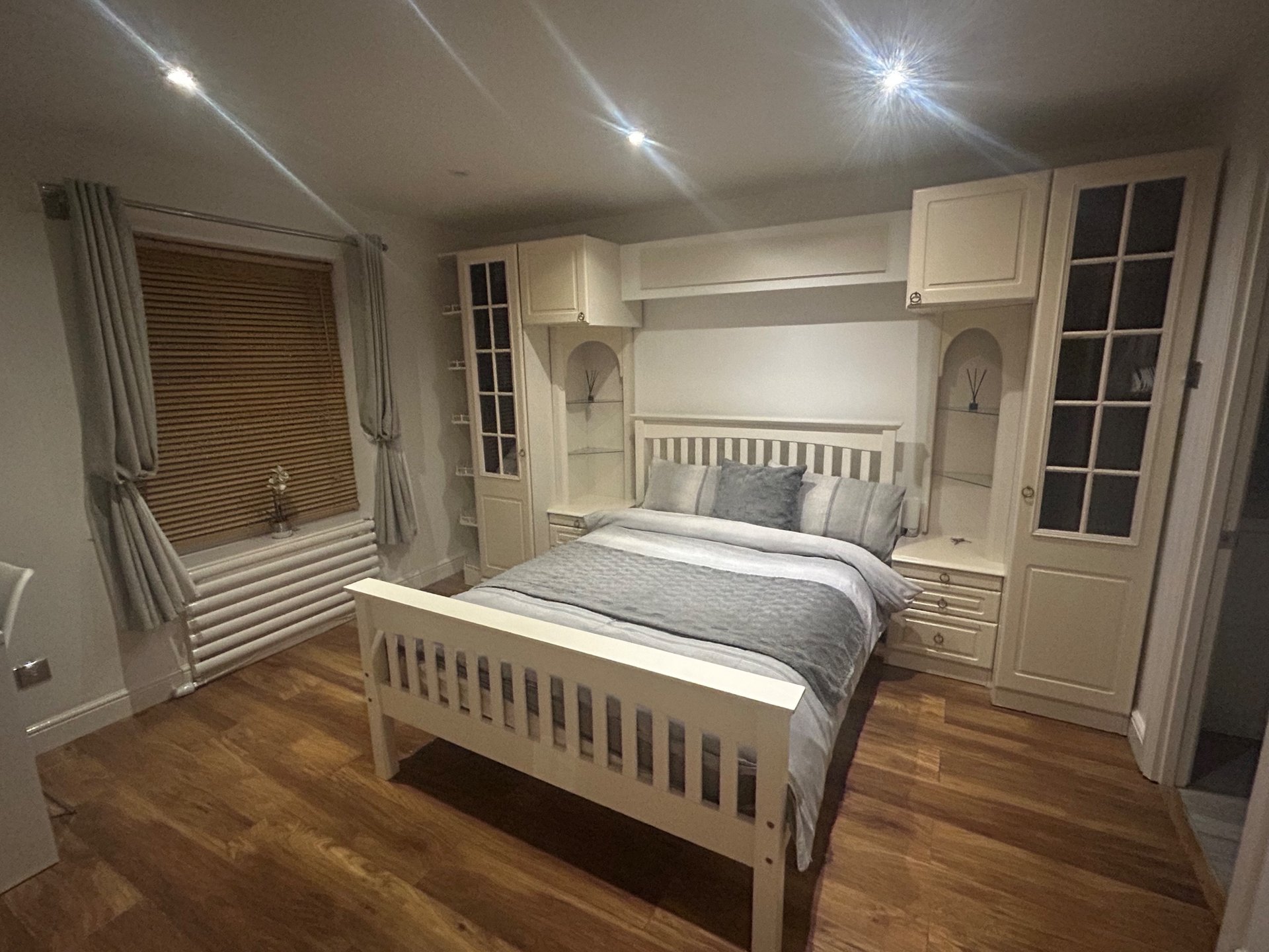 Bright bedroom with white wooden bed, built-in wardrobes, wooden flooring and curtained window