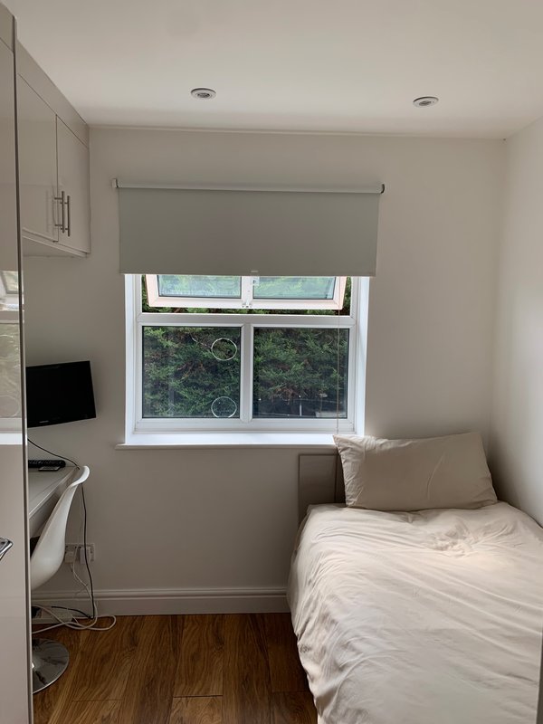Compact bedroom with single bed, desk with monitor, window with blinds and wooden flooring