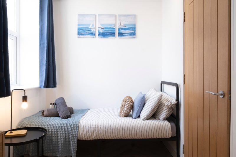 Compact bedroom with single bed, blue striped bedding, cushions, wooden door and coastal wall art