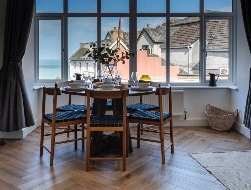 Dining area with wooden table, blue cushioned chairs, and large window overlooking coastal town rooftops