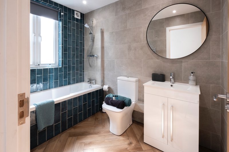 Modern bathroom with teal tile shower, white vanity, round mirror and wooden flooring