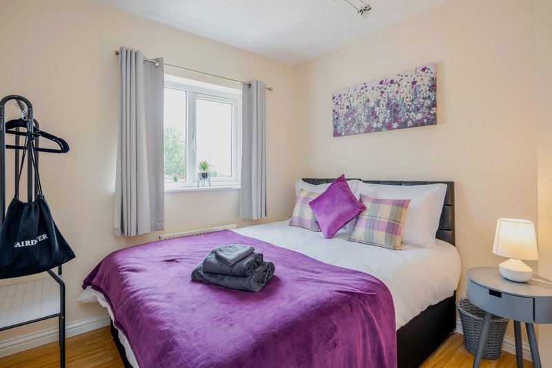 Bright bedroom with purple bedding, double bed, window with curtains, and bedside lamp