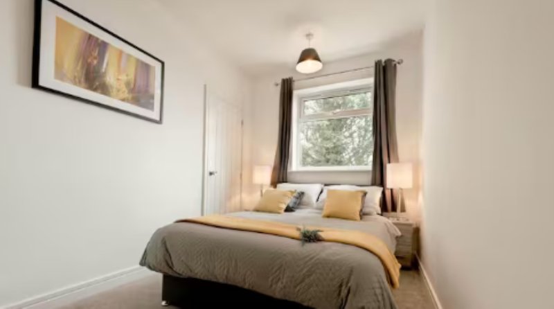 Bright bedroom with double bed, grey bedding, yellow accents, and window with curtains