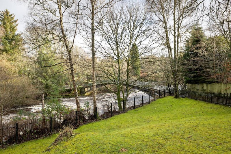 Large garden with green lawn, mature trees, and stream with wooden bridge visible
