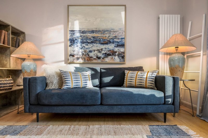 Modern living room with dark blue sofa, striped cushions, table lamps, framed coastal artwork and shelving
