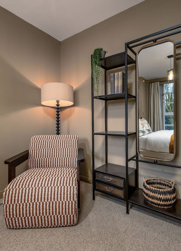 Modern bedroom corner with striped ottoman, table lamp, metal shelving unit and large mirror