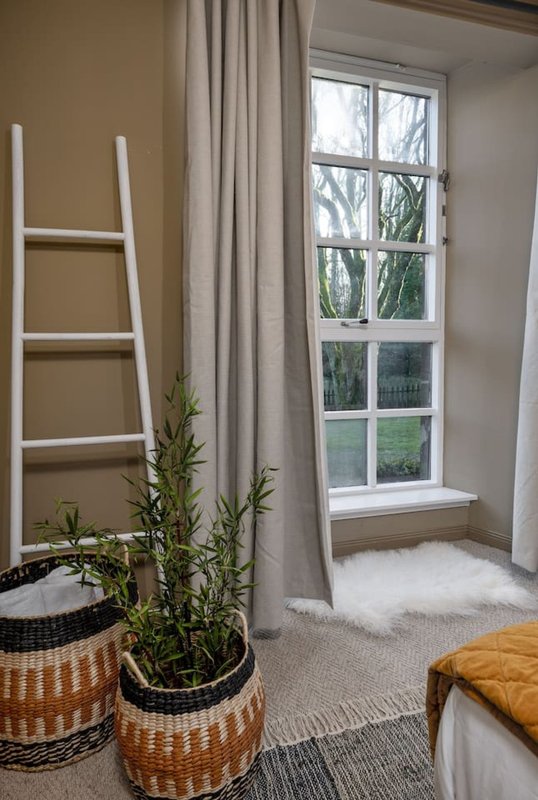 Bright bedroom nook with tall window, cream curtains, decorative ladder, potted plants and cushioned seating