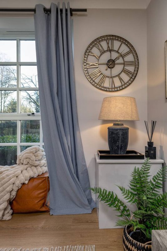 Bedroom corner with window, grey curtains, bed with cushions, table lamp and decorative clock