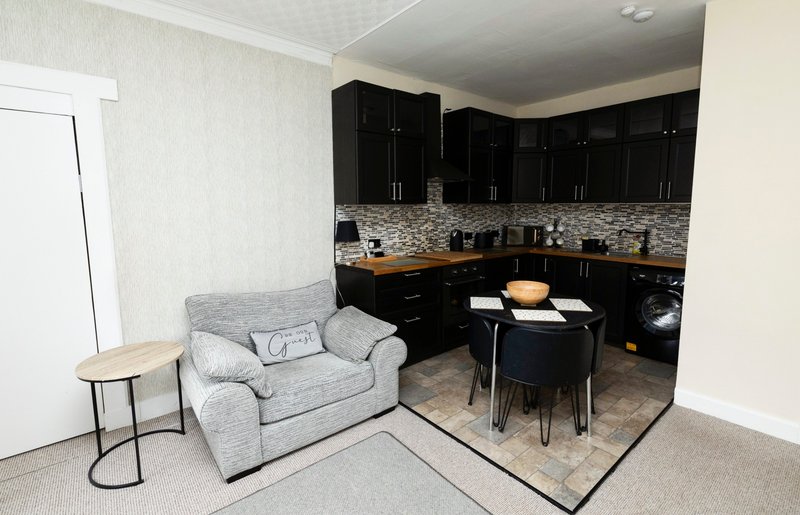 Open-plan living and kitchen area with grey armchair, dining table, black cabinetry and mosaic backsplash