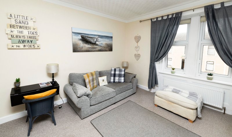 Light living room with grey sofa, decorative cushions, side table lamp, and window with dark curtains