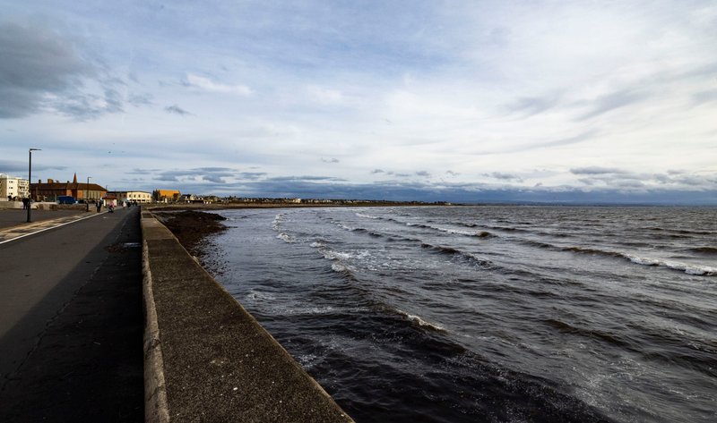 Coastal promenade with sea wall, buildings, and waves under cloudy sky