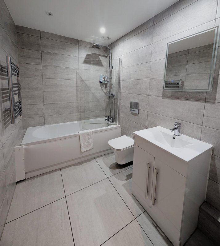 Modern bathroom with grey tile walls, white vanity unit, toilet and bathtub with shower enclosure