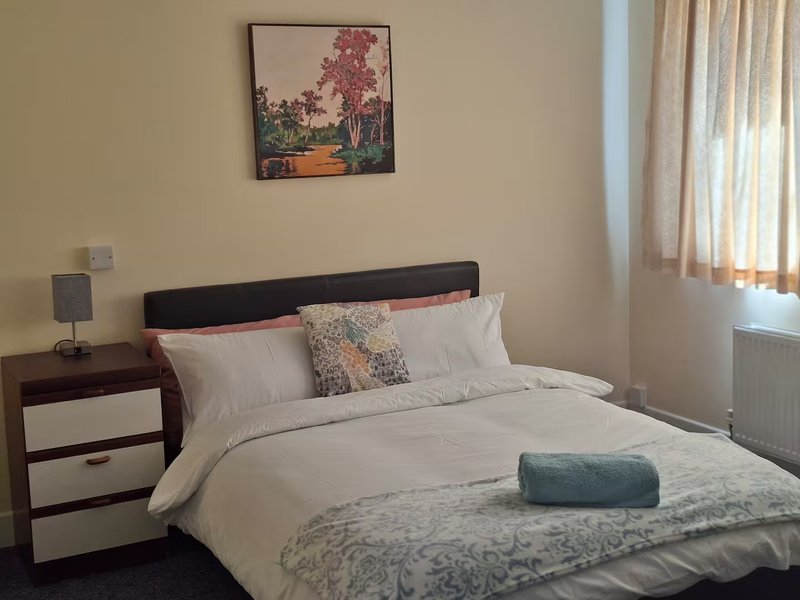 Bedroom with double bed, cream bedding, wooden bedside table, framed artwork and window with curtains