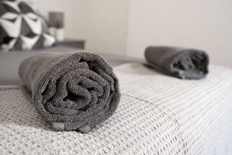 Rolled gray towels on white bedspread with pillows in background
