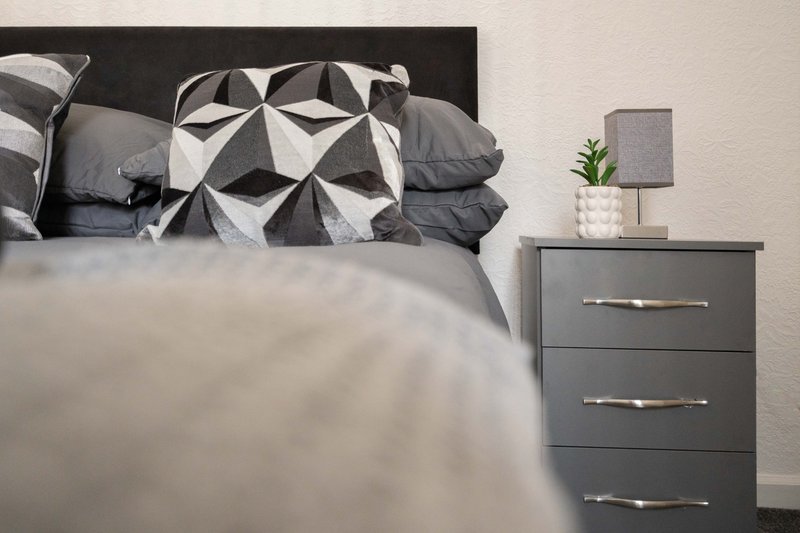 Modern bedroom with grey upholstered bed, geometric patterned cushions, and bedside table with plant.