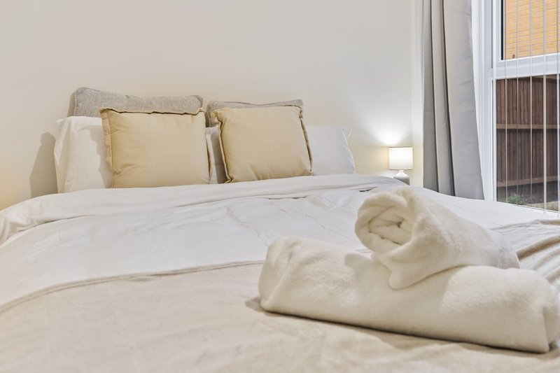 Bright bedroom with cream bedding, decorative pillows, folded towels, and window with blinds