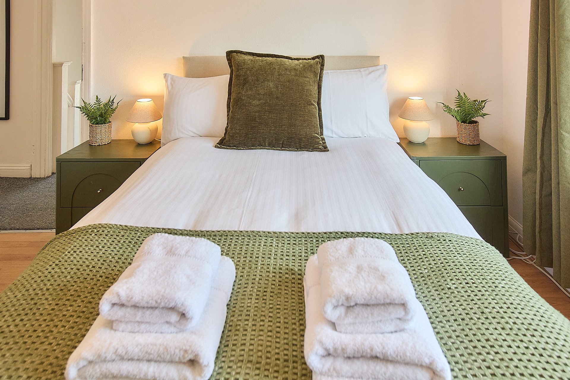 Bedroom with double bed, white bedding, green cushion, nightstands with lamps and plants