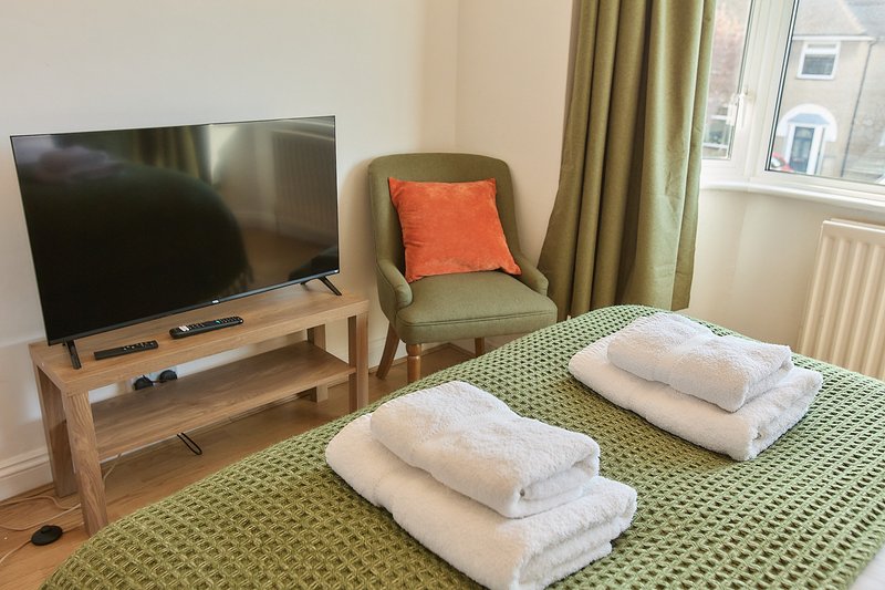 Bedroom with double bed, green patterned bedding, white towels, TV on wooden stand, armchair with orange cushion, and window with green curtains