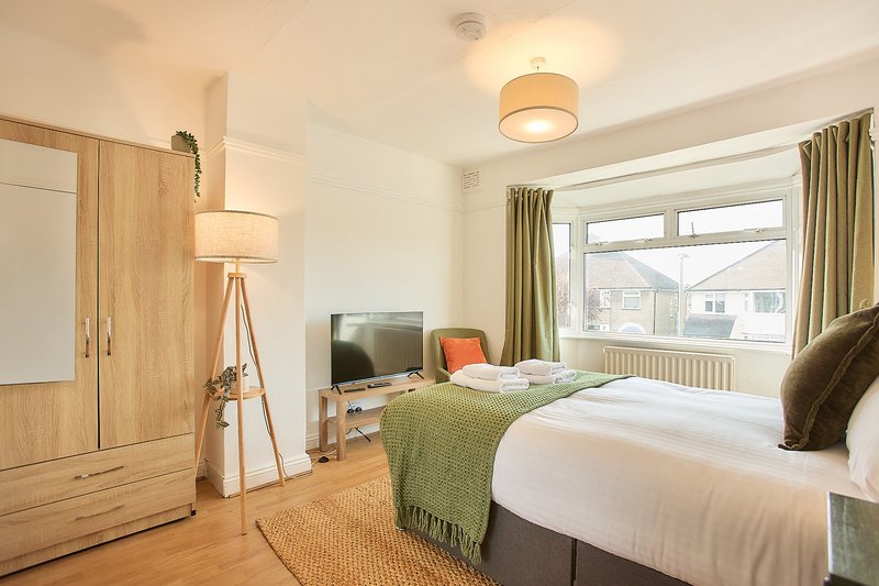 Bright bedroom with double bed, wooden wardrobe, tripod lamp, and views across neighbourhood