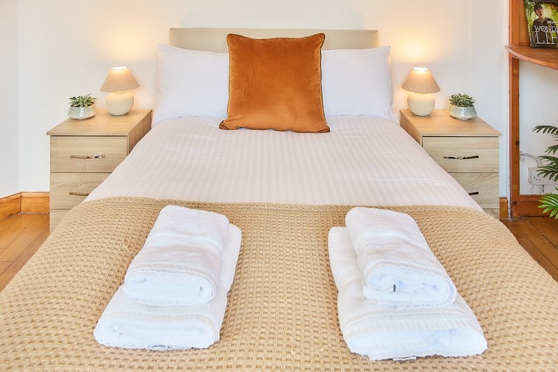 Bright bedroom with double bed, orange cushion, white pillows, wooden nightstands with lamps, and rolled towels on carpet