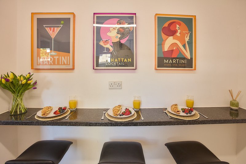 Modern kitchen counter with three place settings, flower vase, juice glasses, and three framed vintage cocktail posters on wall