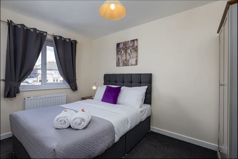 Bedroom with double bed, purple cushion, rolled towels, black headboard and dark curtains