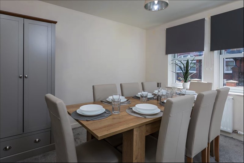 Dining room with wooden table, beige upholstered chairs, place settings, and windows with roller blinds