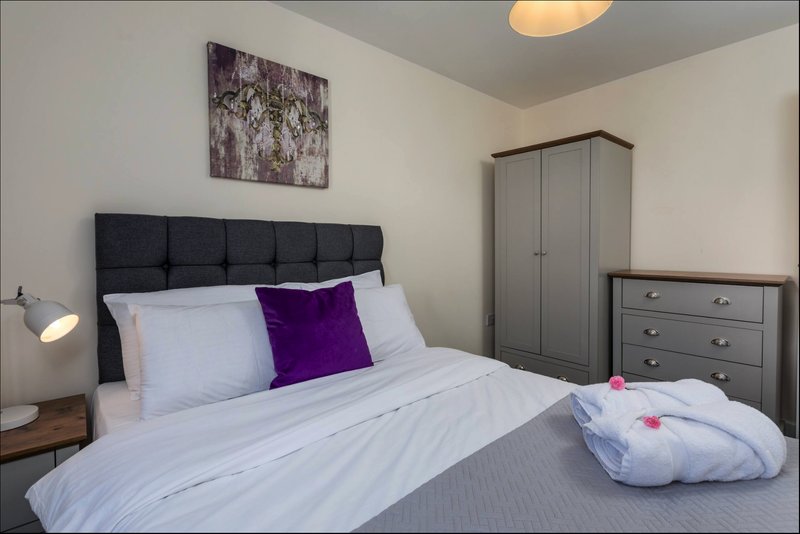 Modern bedroom with double bed, purple and white pillows, dark headboard, and grey furniture